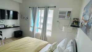 Una habitación de hotel con cama, televisor montado en la pared, escritorio con silla y ventana con cortinas.