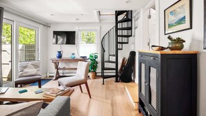 A modern living room with a wooden floor, a sofa, a coffee table, a TV, a staircase, and a fireplace. - Charming Getaway 1 Block from Beach! (Provincetown)