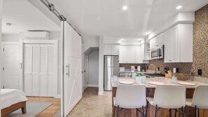 A modern kitchen with white cabinetry, a central island with chairs, a microwave, and a refrigerator. - Charming Getaway 1 Block from Beach! (Provincetown)