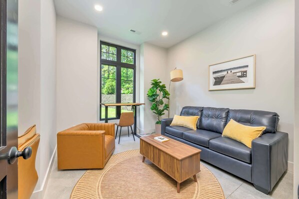 Una sala moderna con un sofá de cuero, una mesa de centro de madera y un sillón amarillo. Se ve una ventana grande con vistas a follaje.