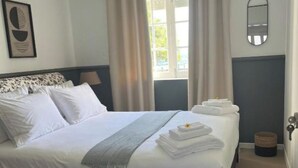 A neatly made bed with white linens and a grey throw, a bedside table, a patterned rug, and a window with curtains. - Two Bedroom Apartment R C Porto Martins Pools (Porto Martins)