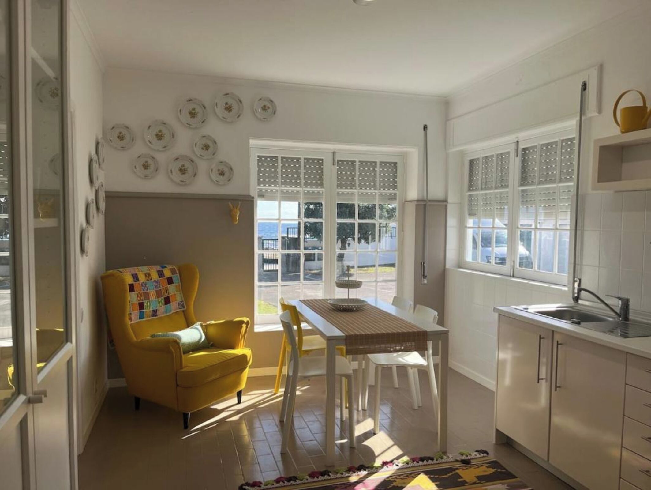 A bright kitchen with a dining area, a yellow armchair, and a view through the window.