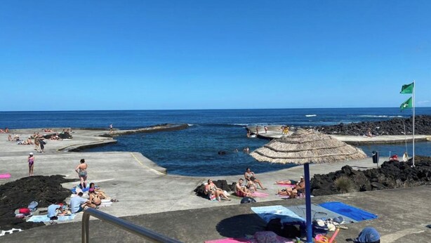 A beach with people sunbathing and swimming, a thatched umbrella, and a flag. - Two Bedroom Apartment R C Porto Martins Pools (Porto Martins)