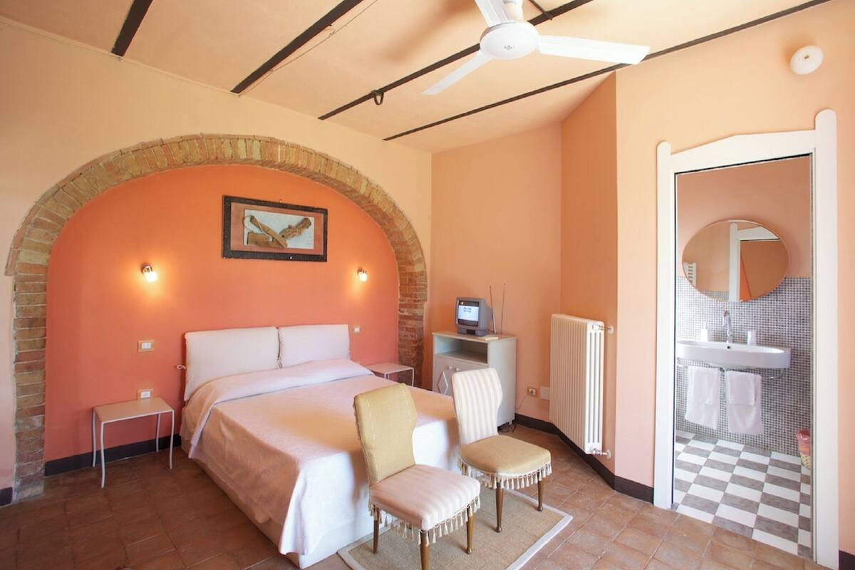 Une chambre à coucher comprenant un lit, une chaise, un bureau, un ordinateur, un radiateur et une salle de bain avec un carrelage à motifs géométriques.
