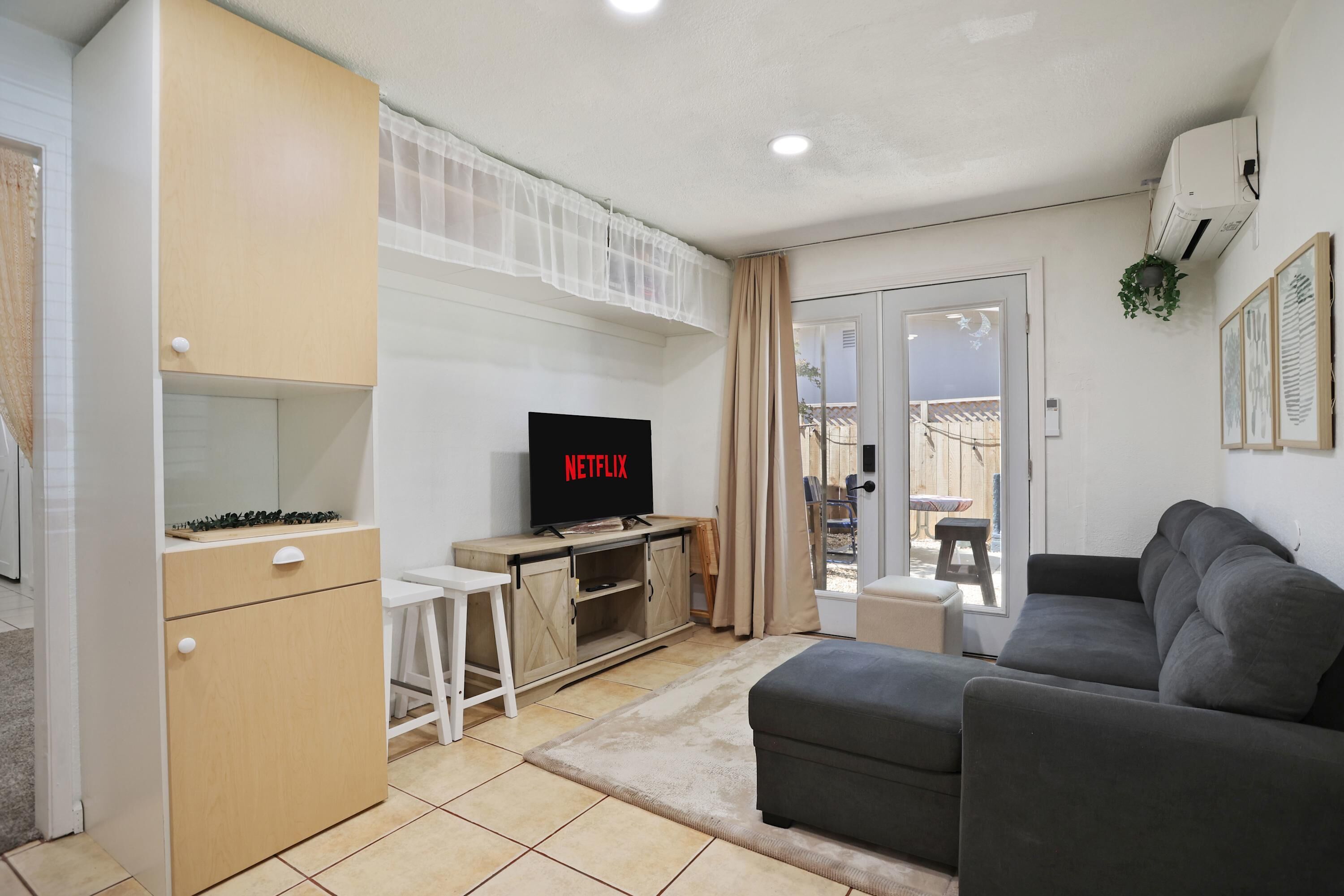 A living room with a grey sofa, a TV displaying 'NETFLIX', a wooden cabinet, and a small white table.