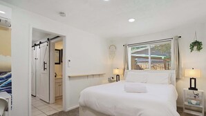 Ein Schlafzimmer mit einem großen Bett, Nachttischlampen, einem Fenster mit Vorhängen, einer hängenden Pflanze und Blick ins Freie.