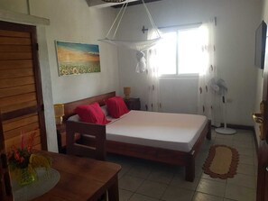 A bedroom with a bed, a wooden table, a painting, a window, and a fan.