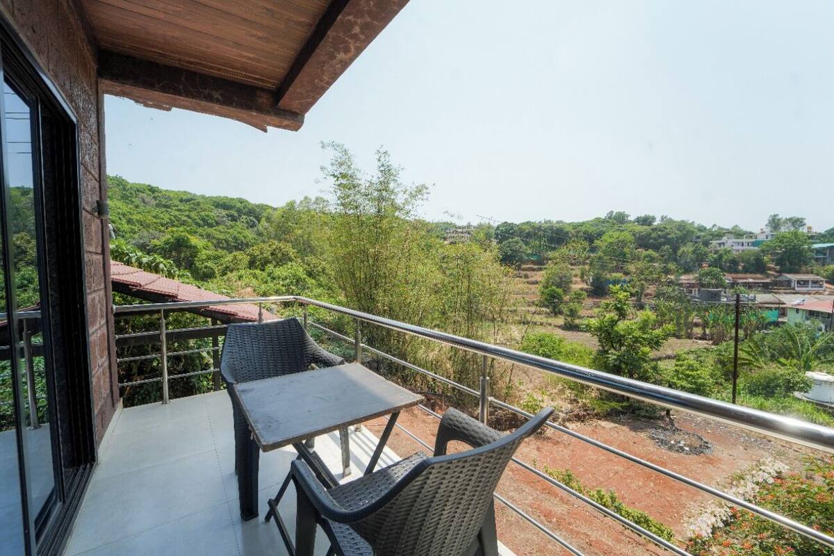 A balcony with a table and chairs, offering a view of a green landscape and distant buildings.