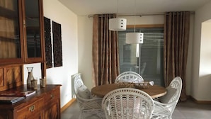 Un comedor con una mesa de madera y sillas de mimbre blanco, un armario de madera y un ventanal con cortinas.