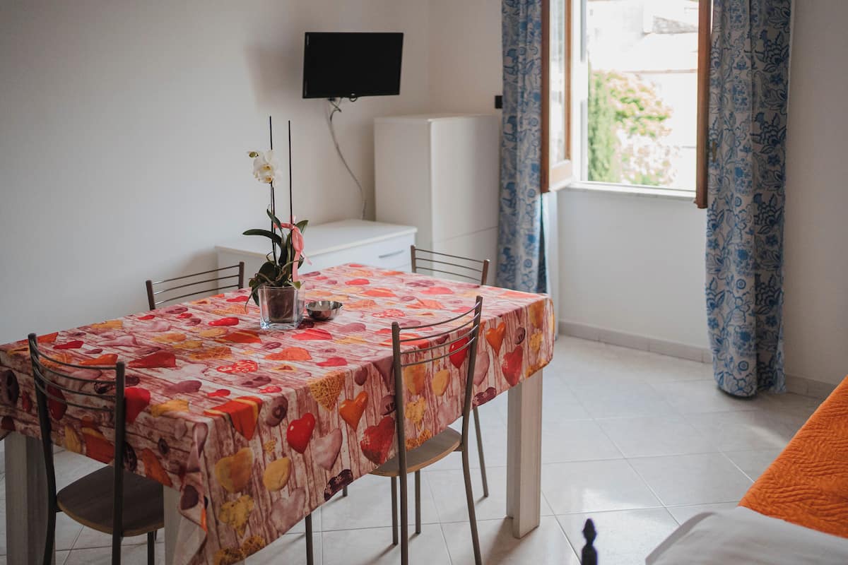 A dining area with a table covered in a floral tablecloth, a potted plant, and a television mounted on the wall.