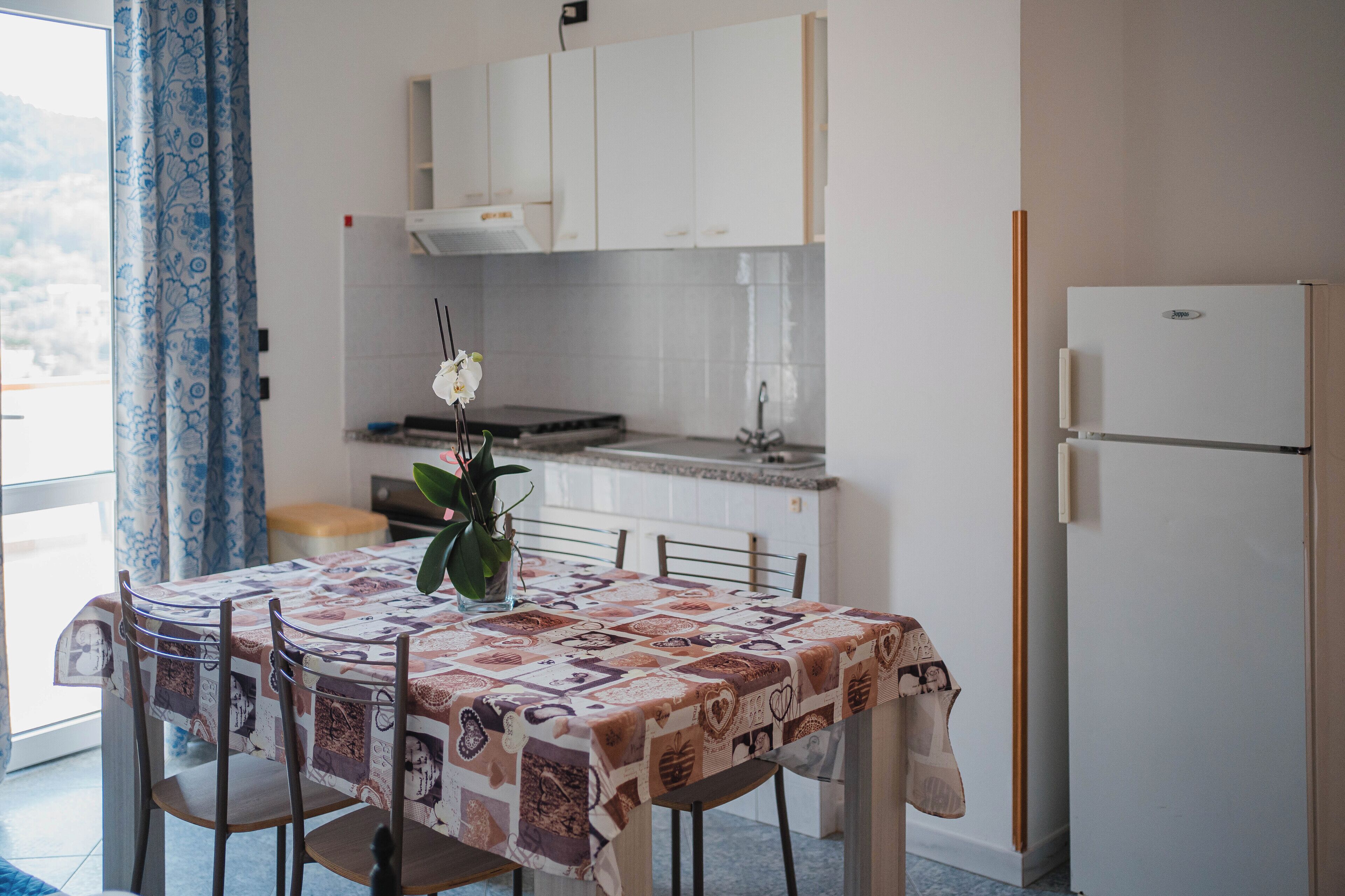 Een kleine keuken met witte kastjes, een koelkast en een eettafel met stoelen.