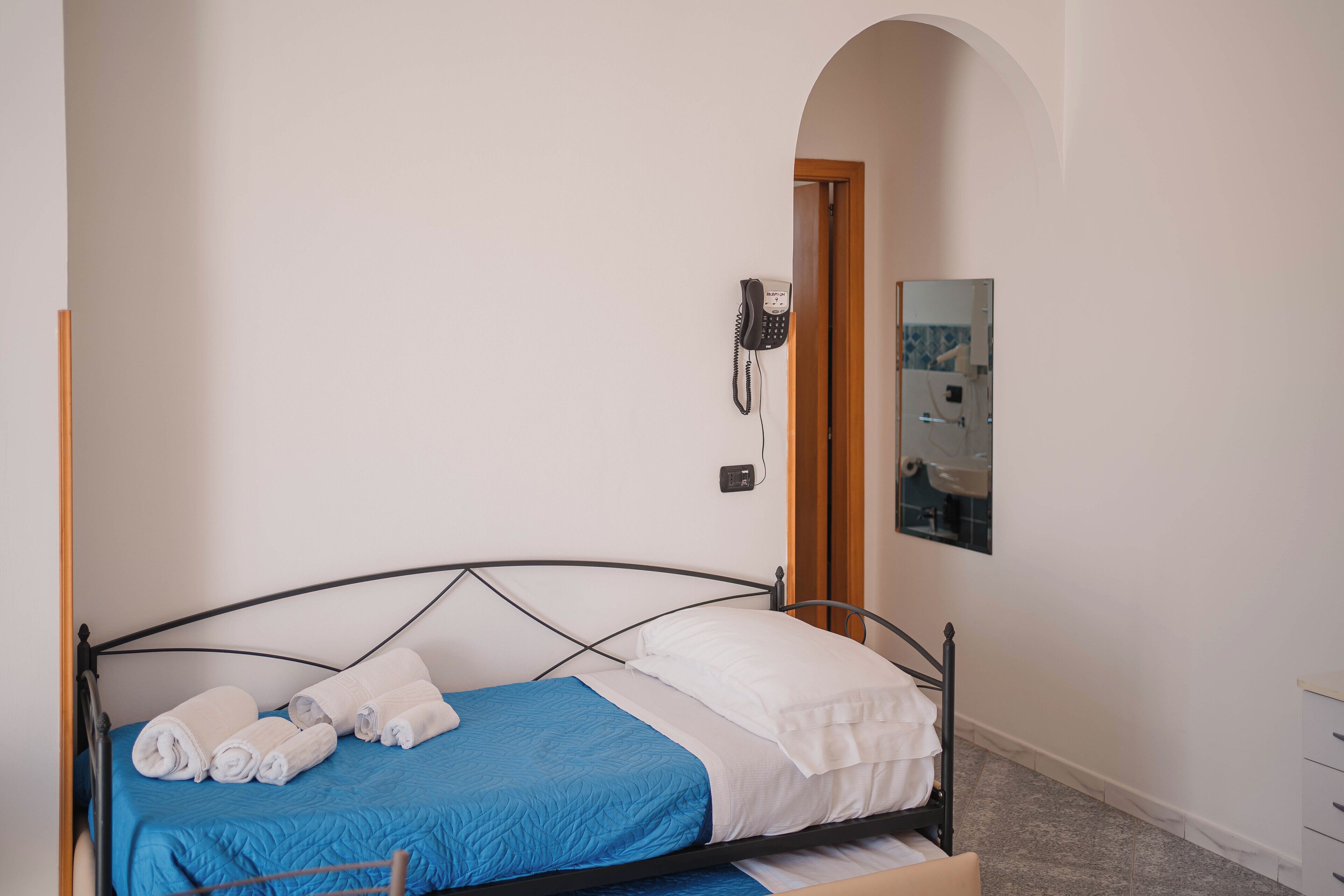 Un letto rifatto con cura, coperto da lenzuola bianche e azzurre, un telefono sulla parete e uno specchio che riflette parte della stanza.