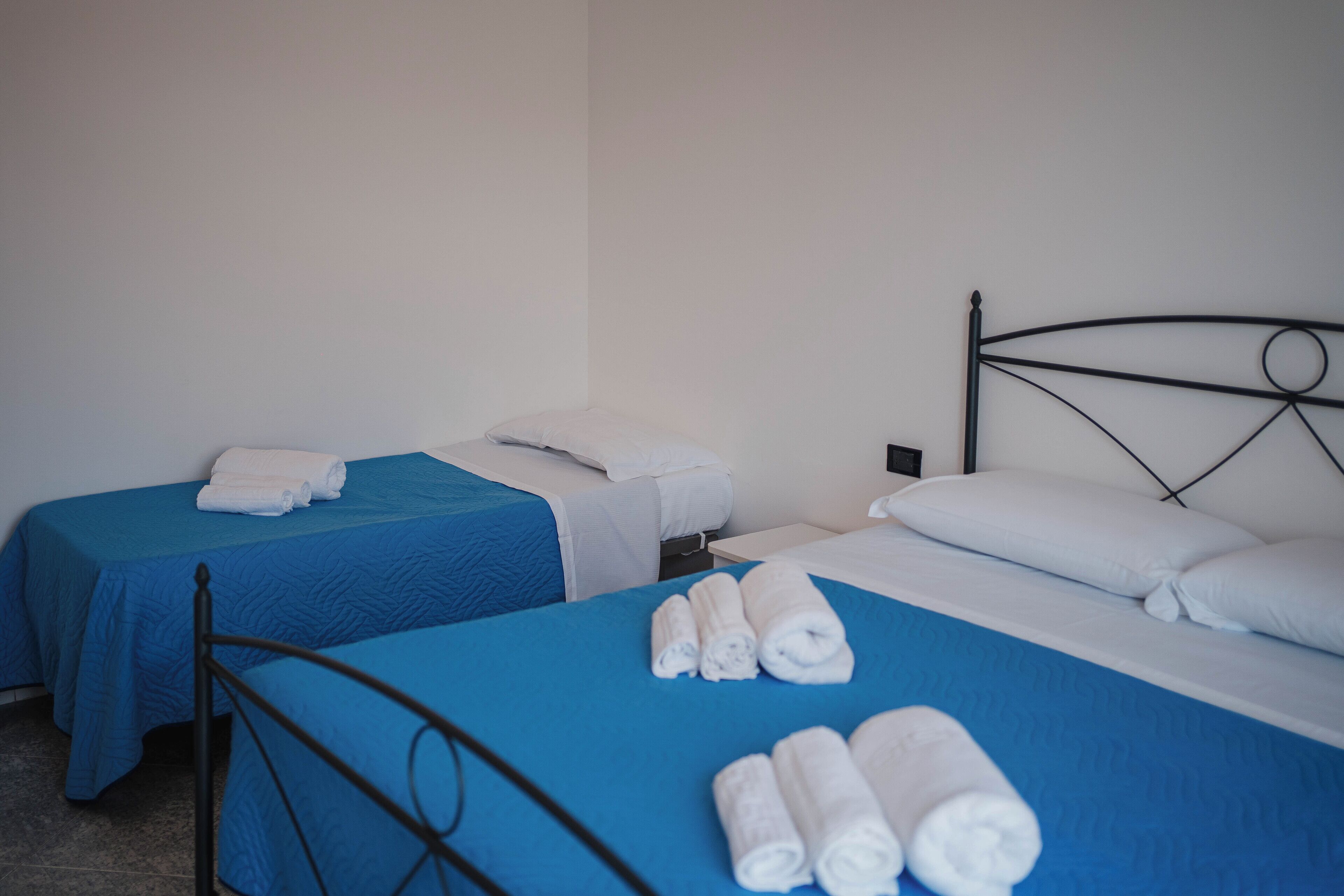 Una camera d'albergo con due letti singoli, ciascuno con copriletto blu e cuscini bianchi. Sui letti sono presenti asciugamani arrotolati.
