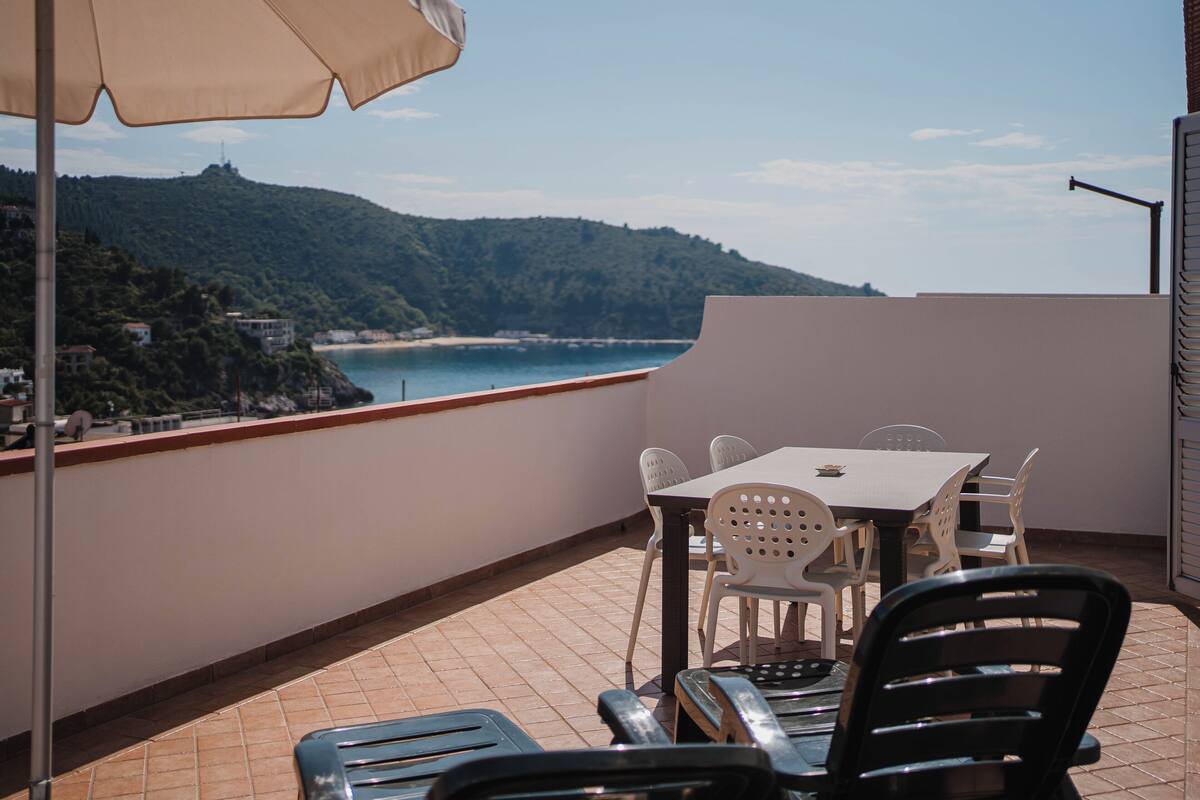 A balcony with a table and chairs, overlooking a coastal landscape with a view of the sea and hills.