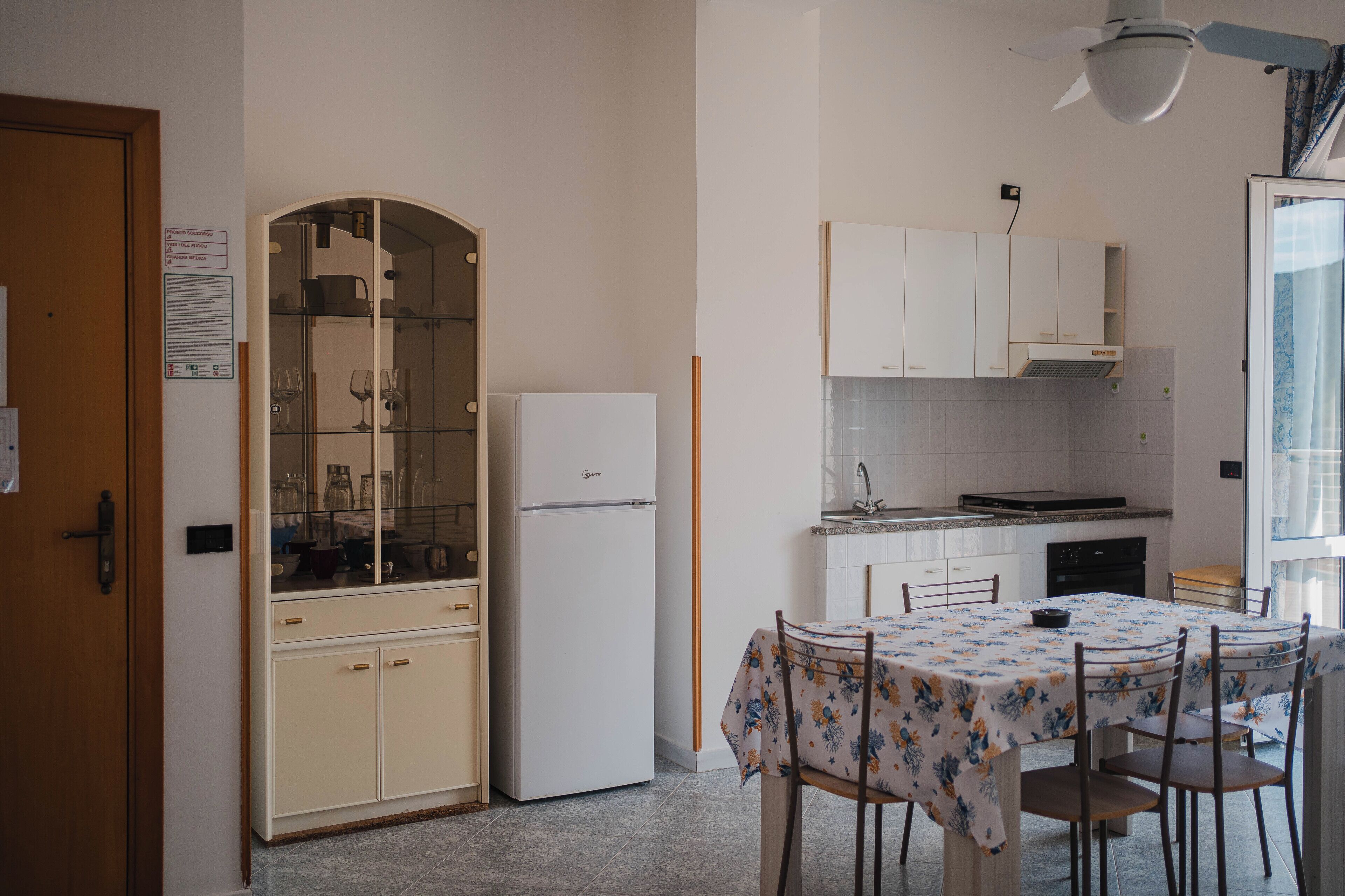 A compact kitchen with a refrigerator, a dining table with chairs, and a glass cabinet with wine glasses.