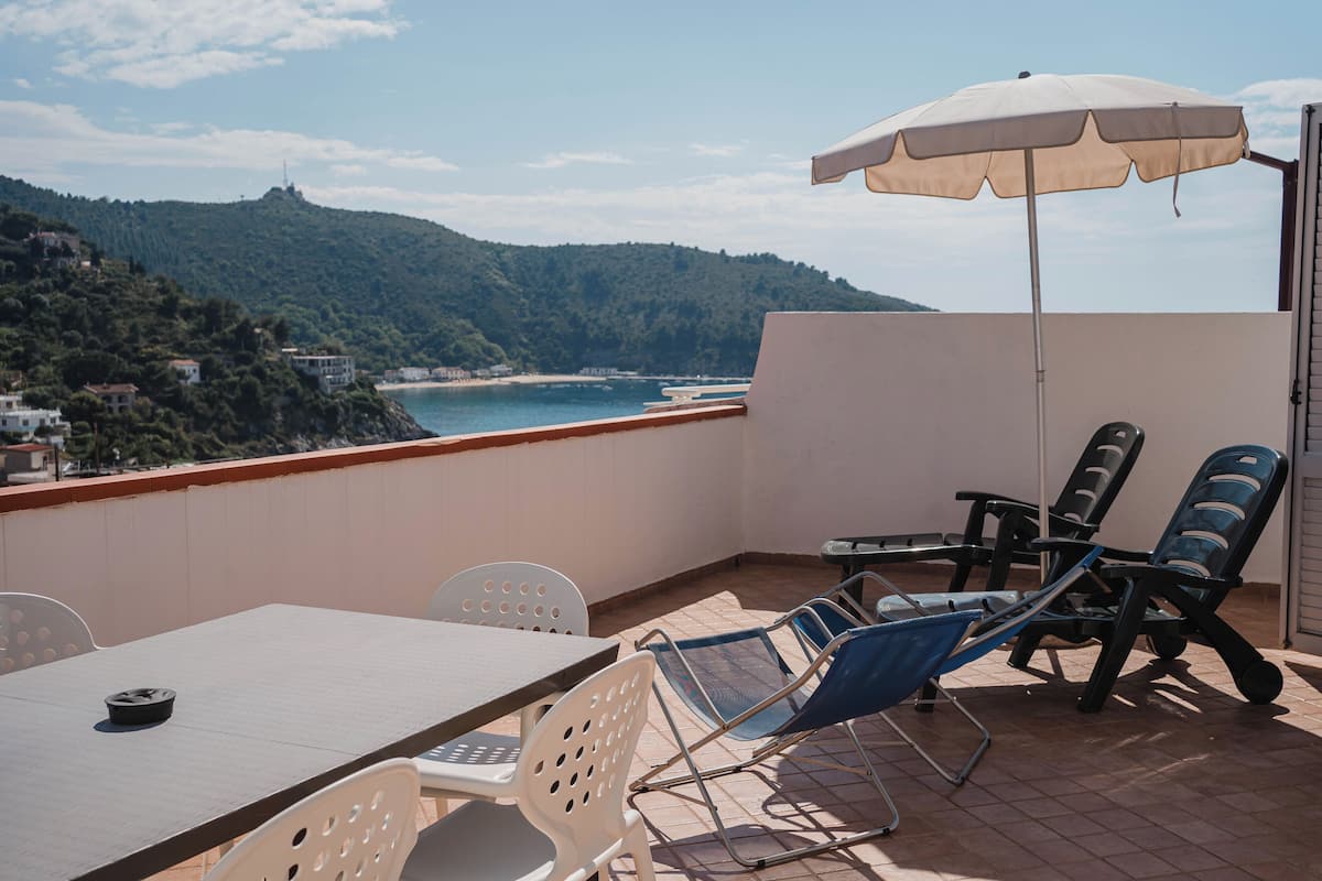 A balcony with a table, chairs, and an umbrella overlooking a coastal view with hills in the background.