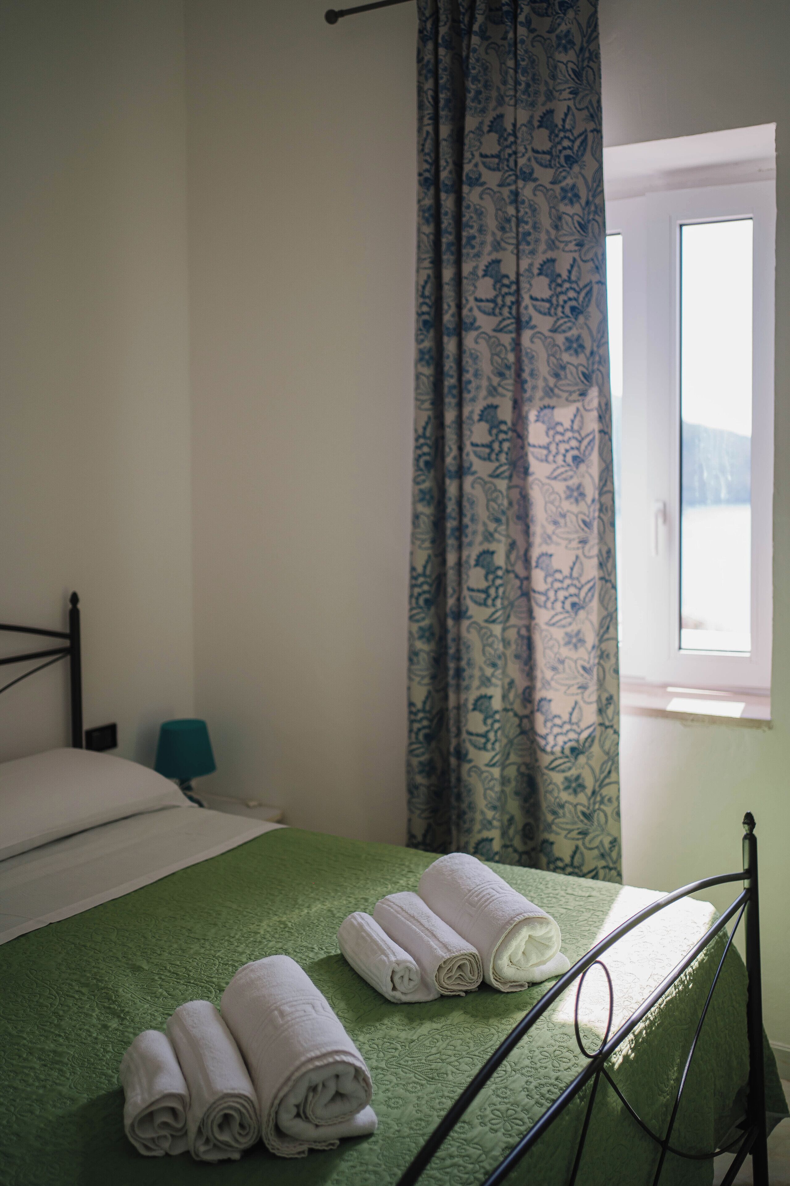 Ein ordentlich gemachtes Bett mit grüner Bettwäsche und drei zusammengerollten Handtüchern, ein Fenster mit Blick und ein blau gemustertes Vorhang.