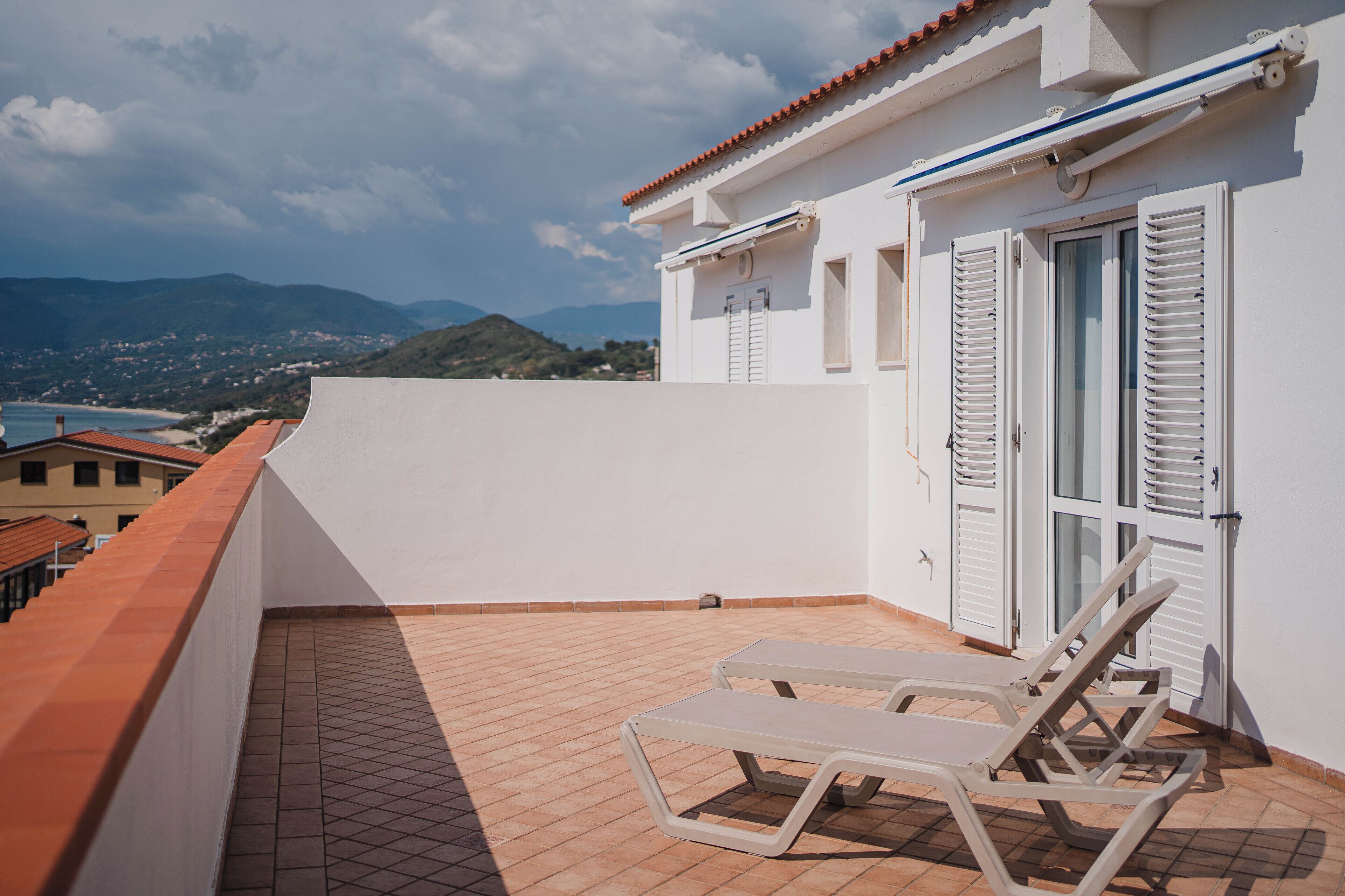 A rooftop terrace with two lounge chairs, a white building with shutters, and a view of distant hills and a beach.