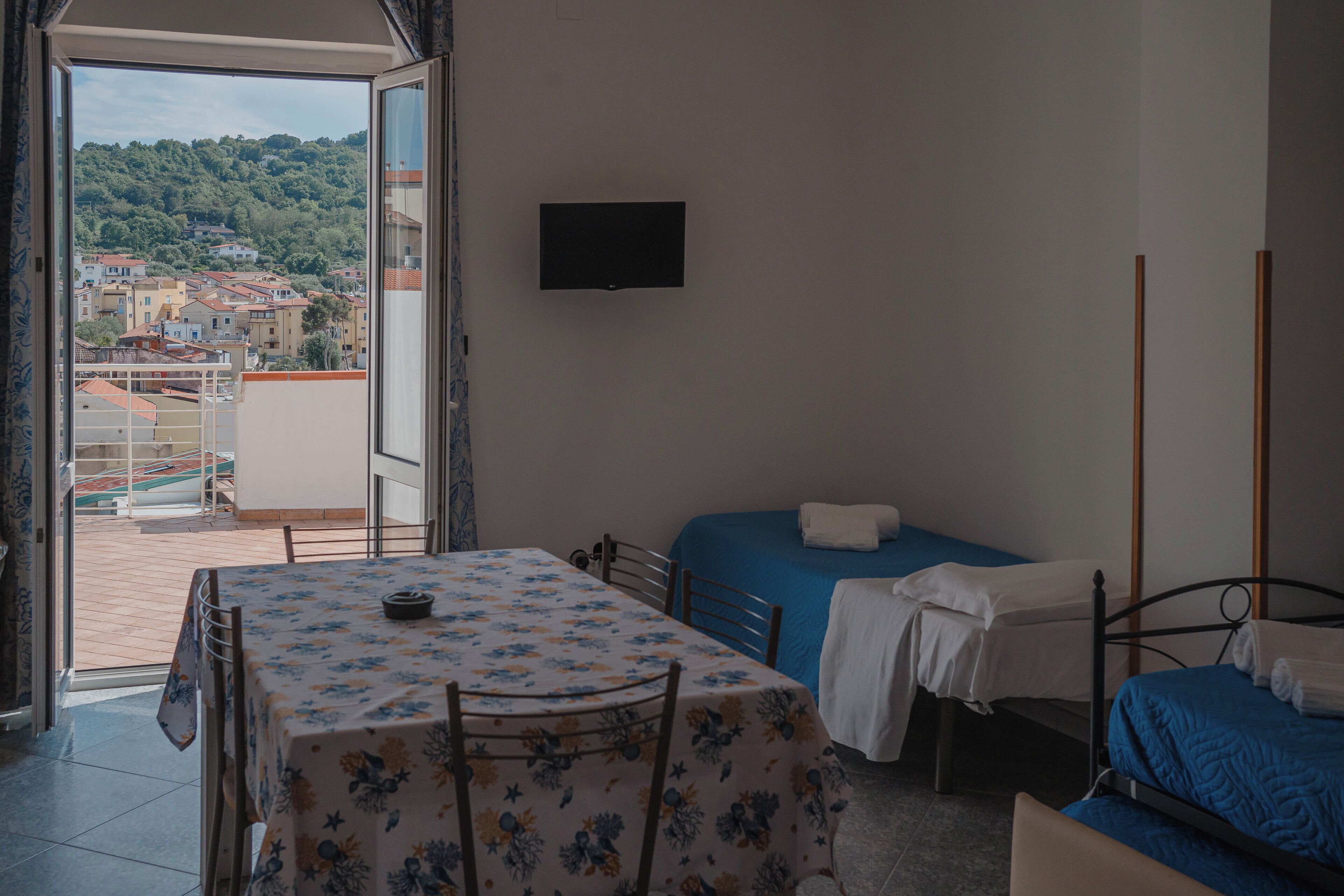 Una camera da letto con balcone, un letto, un tavolo con tovaglia floreale e una TV a parete.