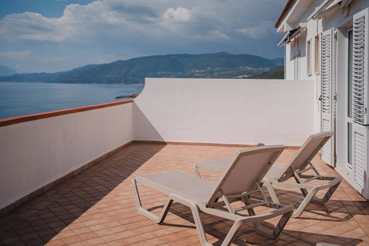 A balcony with two white lounge chairs, a tiled floor, and a view of the sea and mountains.