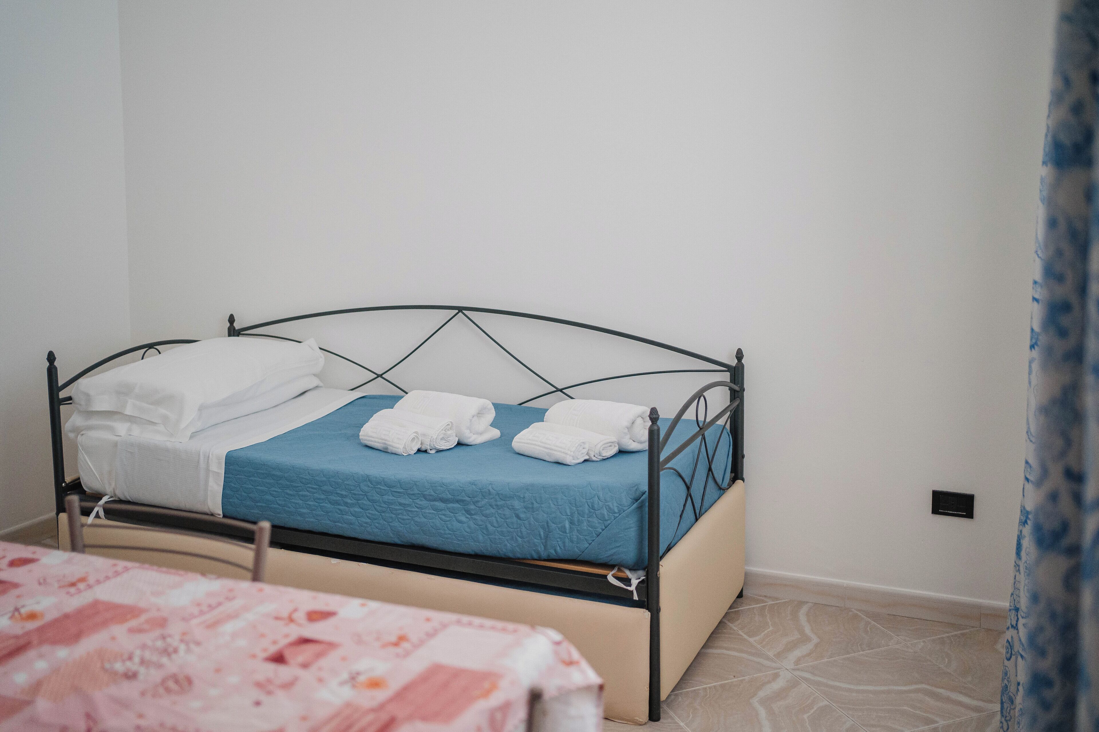 Una cama individual con colchón azul y ropa de cama blanca, una mesita de noche con mantel rosa estampado y una pared blanca lisa con un enchufe.