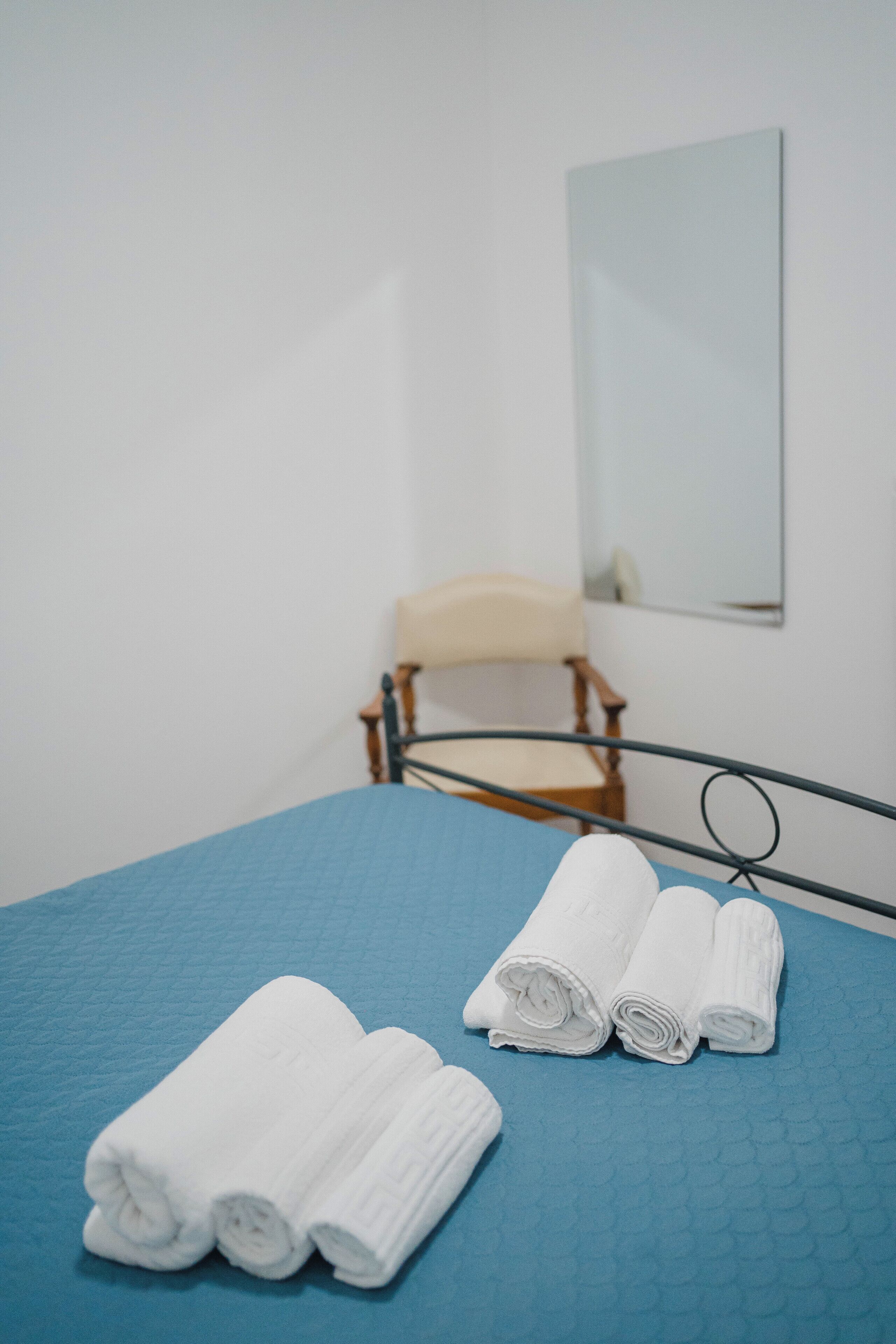 Una cama bien tendida con ropa de cama azul y dos toallas blancas enrolladas. Una silla de madera está colocada contra la pared, y un espejo cuelga encima.