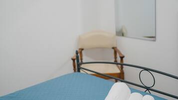 Una cama bien tendida con ropa de cama azul y dos toallas blancas enrolladas. Una silla de madera está colocada contra la pared, y un espejo cuelga encima.