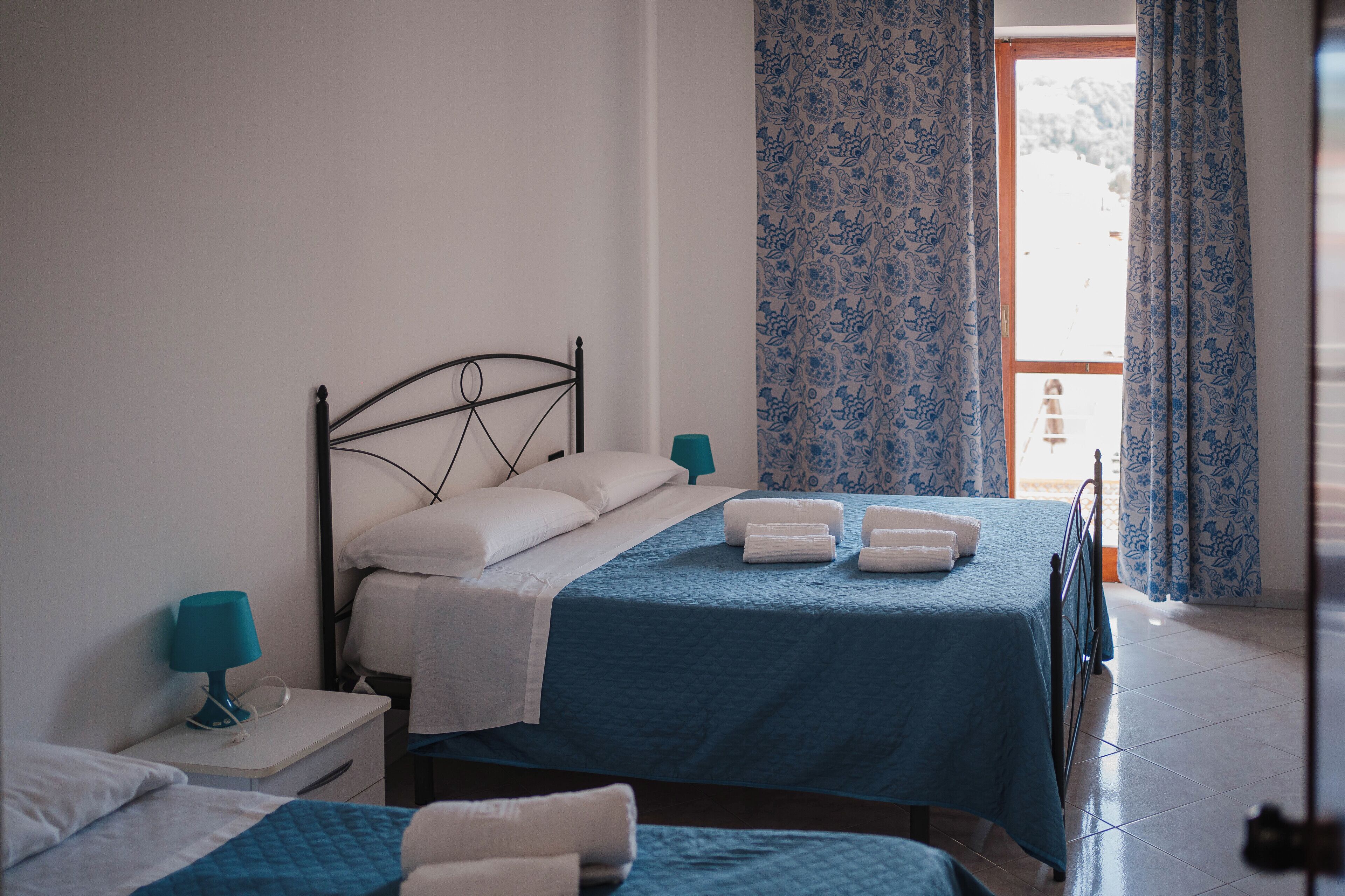 A bedroom with two beds, a nightstand with a lamp, and a view of the outdoors through a window with curtains.
