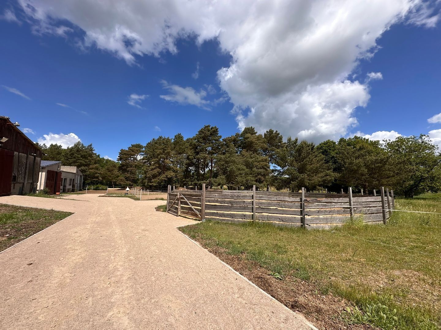 A rural property with a dirt path leading to a wooden gate, surrounded by trees and a clear sky.