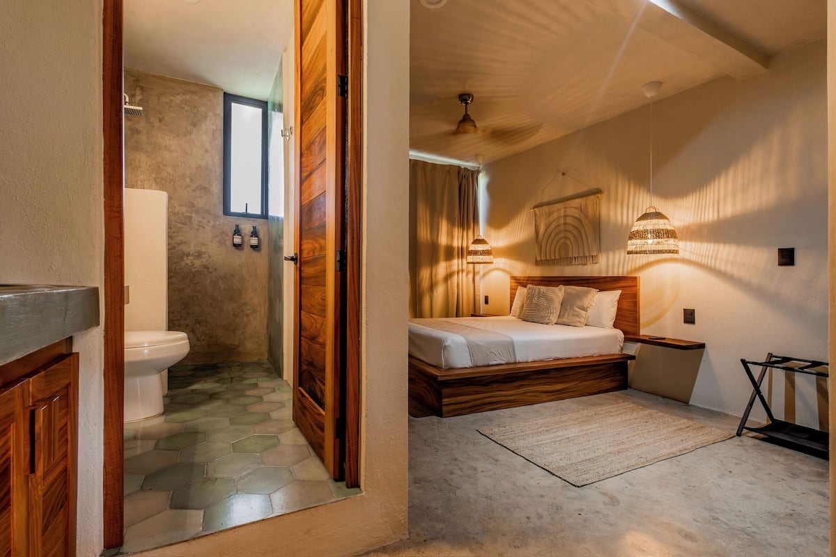 A bedroom with a wooden bed, a bedside table, a hanging light fixture, and a view of the bathroom with a toilet and shower.