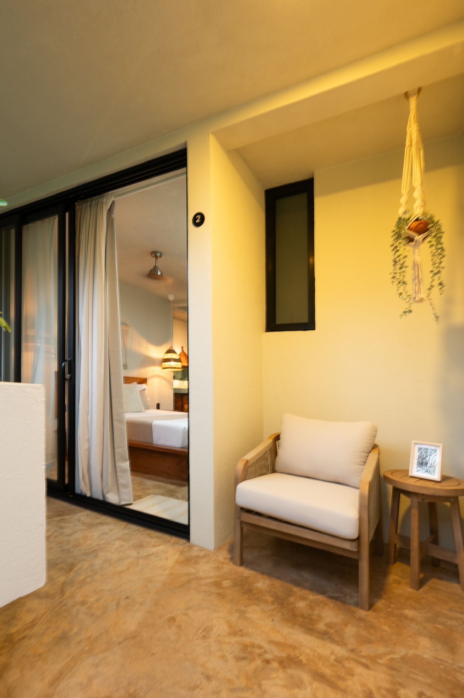 A hotel room with a bed, a chair, a small table, and a hanging plant.
