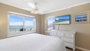 A bedroom with a large bed, a ceiling fan, a TV, and a view of the beach. - Tradewinds 605 by Liquid Life (Orange Beach)