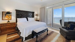 A bedroom with a large bed, a wooden dresser, a bench, a ceiling fan, and a view of the ocean through a sliding door. - Regency Isle 1010 by Liquid Life (Orange Beach)