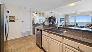 A modern kitchen with a granite countertop, stainless steel appliances, and a view of the ocean through large windows. - Regency Isle 1010 by Liquid Life (Orange Beach)