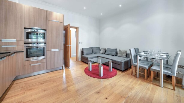 A modern kitchen with built-in ovens, a dining area with a glass table and grey chairs, and a grey sofa. - Contemporary 1B apartment - Paddington St (Greater London)