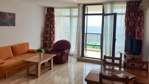 Ein Wohnzimmer mit einer Couch, einem Sessel, einem Tisch und einem Fernseher. Ein großes Fenster mit Vorhängen bietet Blick aufs Meer.