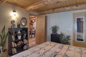 Un dormitorio acogedor con una cama grande, una estantería, una puerta de madera y un pequeño comedor visible a través de un acceso abierto.