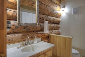 Un baño con pared de tronco de madera, un lavabo blanco, un espejo, una toalla colgada en un perchero y una lámpara.