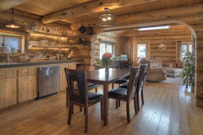 Interior rústico de una cabaña con una mesa de comedor de madera, sillas, una cocina con armarios de madera y una sala de estar acogedora con sofá y chimenea.