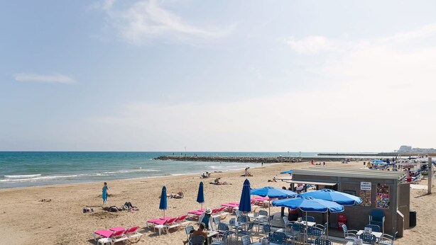 Ein Strand mit Liegestühlen, Sonnenschirmen, einer Mole und einem Rettungsturm.
