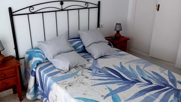 Un lit recouvert d’une couvre-lit à motifs floraux, deux oreillers, une table de chevet avec une lampe, et une porte visible au fond.