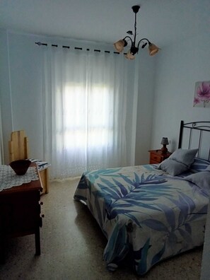 Un dormitorio con cama, mesita de noche, silla de madera, mesa con un bol y ventana con cortinas.
