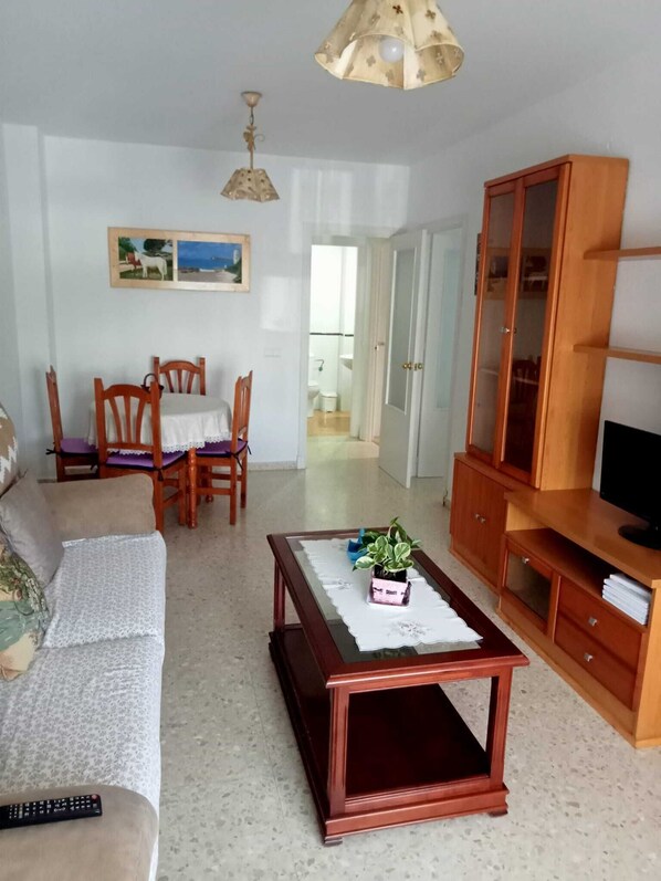A living room with a sofa, a coffee table, a TV stand, and a dining area with chairs and a table.
