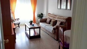 Un salon avec un canapé, une table basse, un fauteuil et une plante. On peut voir des rideaux et une porte.