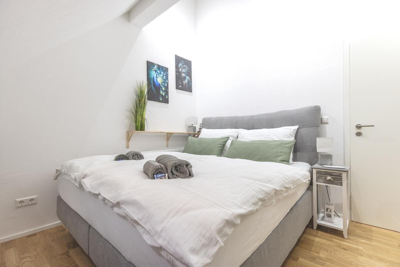 Ein ordentlich bezogenes Bett mit aufgerollten Handtüchern, ein Nachttisch mit Lampe und zwei Bilder darüber.