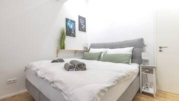 Ein ordentlich bezogenes Bett mit aufgerollten Handtüchern, ein Nachttisch mit Lampe und zwei Bilder darüber.