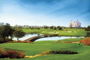 Un parcours de golf avec un pont enjambant un étang, entouré de verdure et avec des bâtiments en arrière-plan.