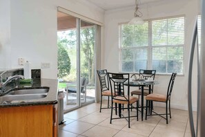 Une cuisine équipée d’un évier, d’une table à manger avec des chaises, d’une fenêtre avec des stores et d’une vue sur la verdure à l’extérieur.