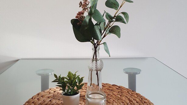 Een ronde tafel met een geweven onderzetter, twee potplanten, een glazen vaas met een kaars en een glazen tafelblad.