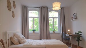 Een slaapkamer met een bed, een bureau en een stoel. Er zijn ramen met gordijnen, een hanglamp en een kamerplant.